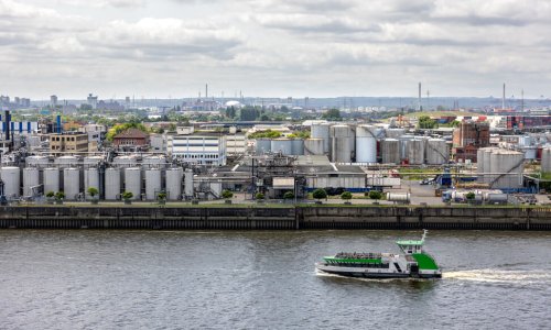 Industrial panorama. River transport. Factory zone. Wide view of an industrial riverbank filled with metal tanks and infrastructure. A small vessel passes by, adding movement to the industrial scene.